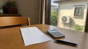 日本の住宅のダイニングテーブルに置かれた電気の検針票と電卓、窓の外に夏の日差しと室外機